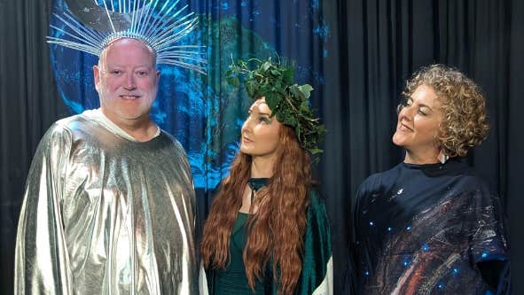 2 women are looking at a smiling man in a gold smock with a silver headpiece
