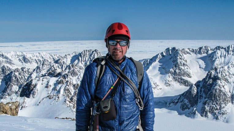 A man in outdoor clothing and mirrored sunglasses is standing against background of snowy mountains.