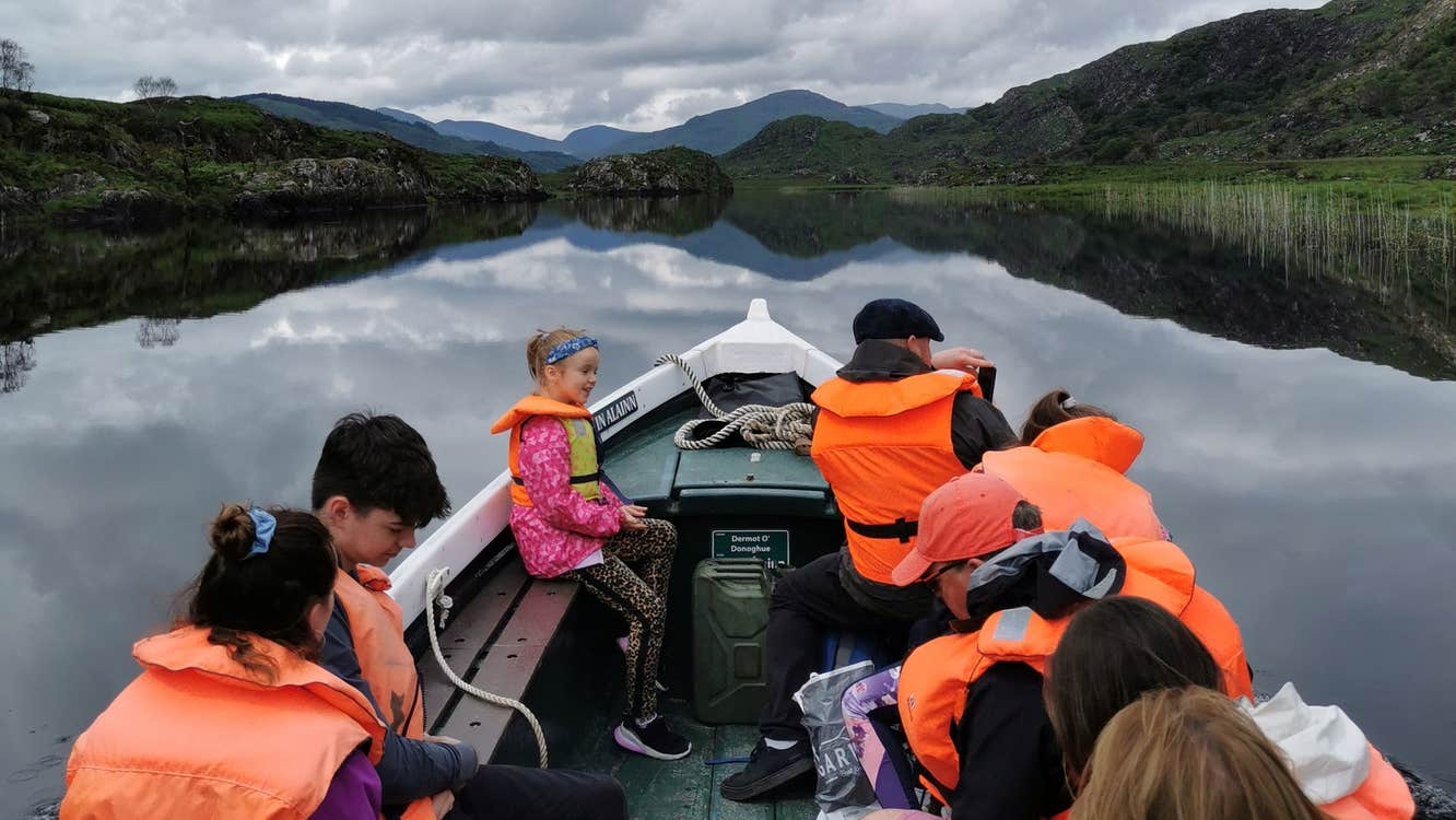 Group of people wearing orange life jackets sitting on a boat sailing on a lake with mountains in the background