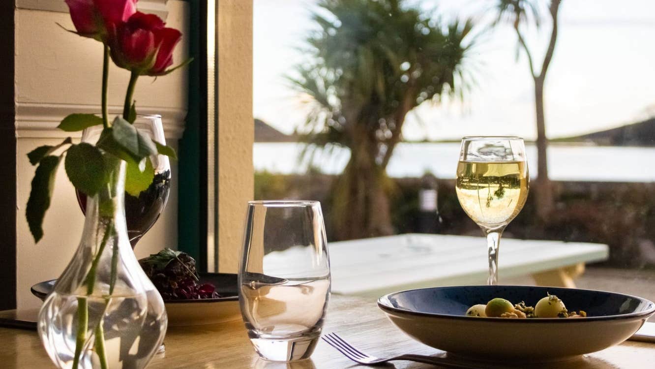 Table with food a rose in a vase and two glasses on it and views of some trees and the sea