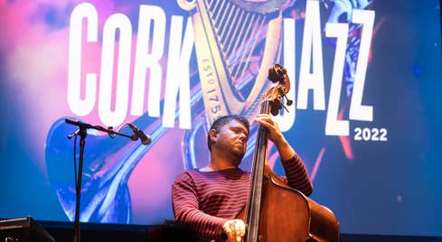GoGo Penguin  at 2022 Guinness Cork Jazz Festival. Person playing a cello or double bass with festival logo on screen behind them.