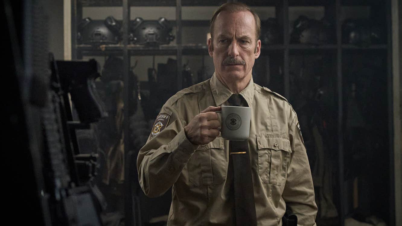 A man wearing a sheriff's uniform is in an armoury. He is holding a mug.