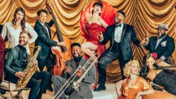 A group of musicians with instruments in vintage suits and dresses, some seated or standing or laying on a light coloured carpet.