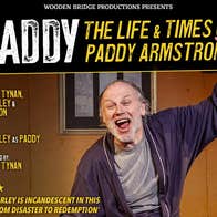 A man in a dressing gown is shouting with arms outstretched on a stage set