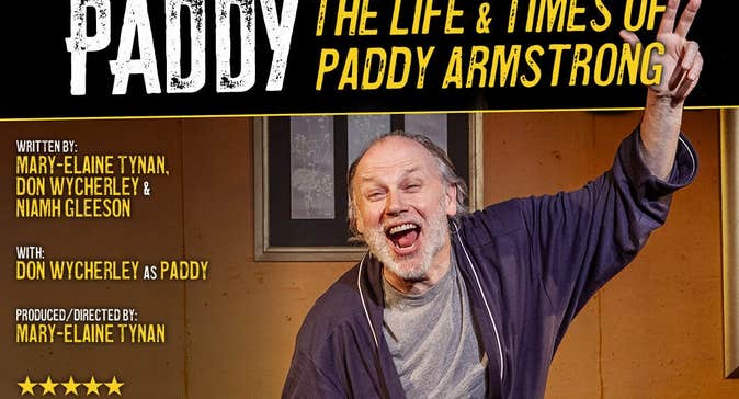 A man in a dressing gown is shouting with arms outstretched on a stage set