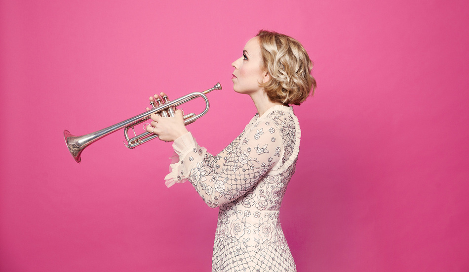 Side view of a woman in pale dress with silver patterns, holding up a trumpet in front of her, against bright pink background.