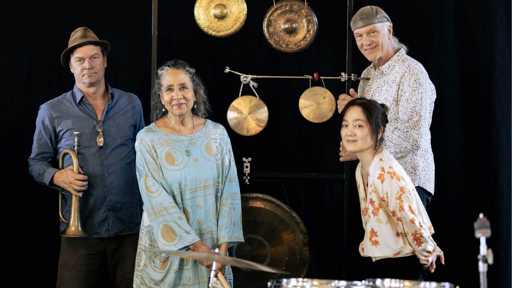 Photo by Patrician Pereira of 4 smiling people with percussion instruments including large drum and various round cymbals hanging on a display against black background.