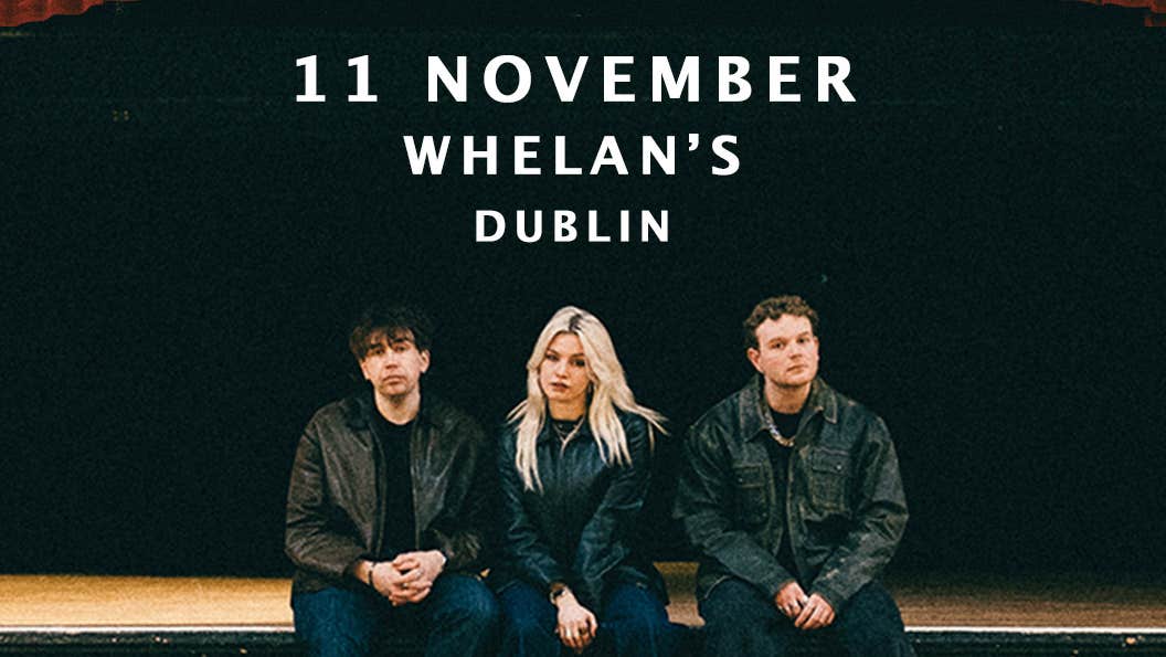 3 people dressed in black clothes and jackets are seated on the edge of a wooden stage.