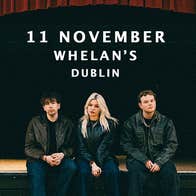 3 people dressed in black clothes and jackets are seated on the edge of a wooden stage.