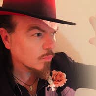 Close up view of man wearing a hat, holding up one hand as if to protect from bright light.