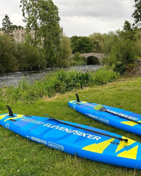 Two blue stand up paddleboards lie flat on a river bank near an old bridge
