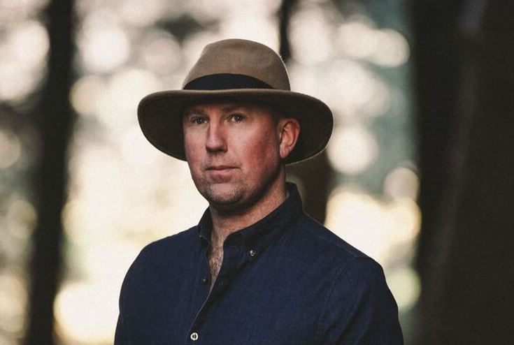 A man in dark shirt wearing a brown hat is outdoors, looking serious.