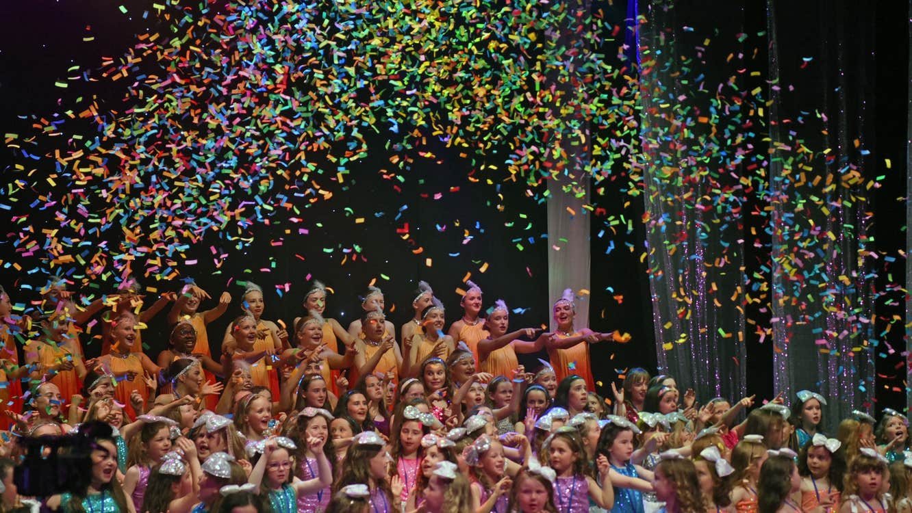 A large group of children gathered on a stage with confetti falling from above