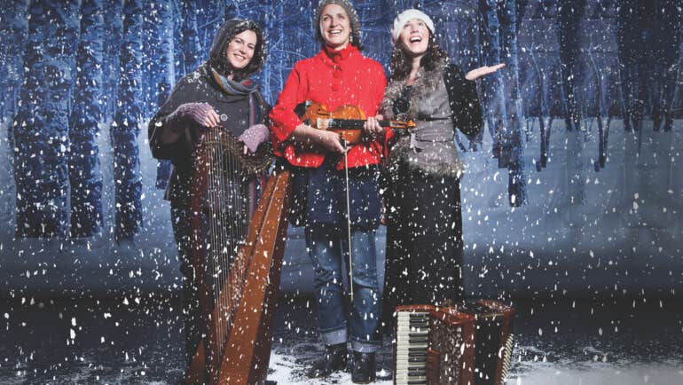 3 smiling women standing with musical instruments in snowy scene