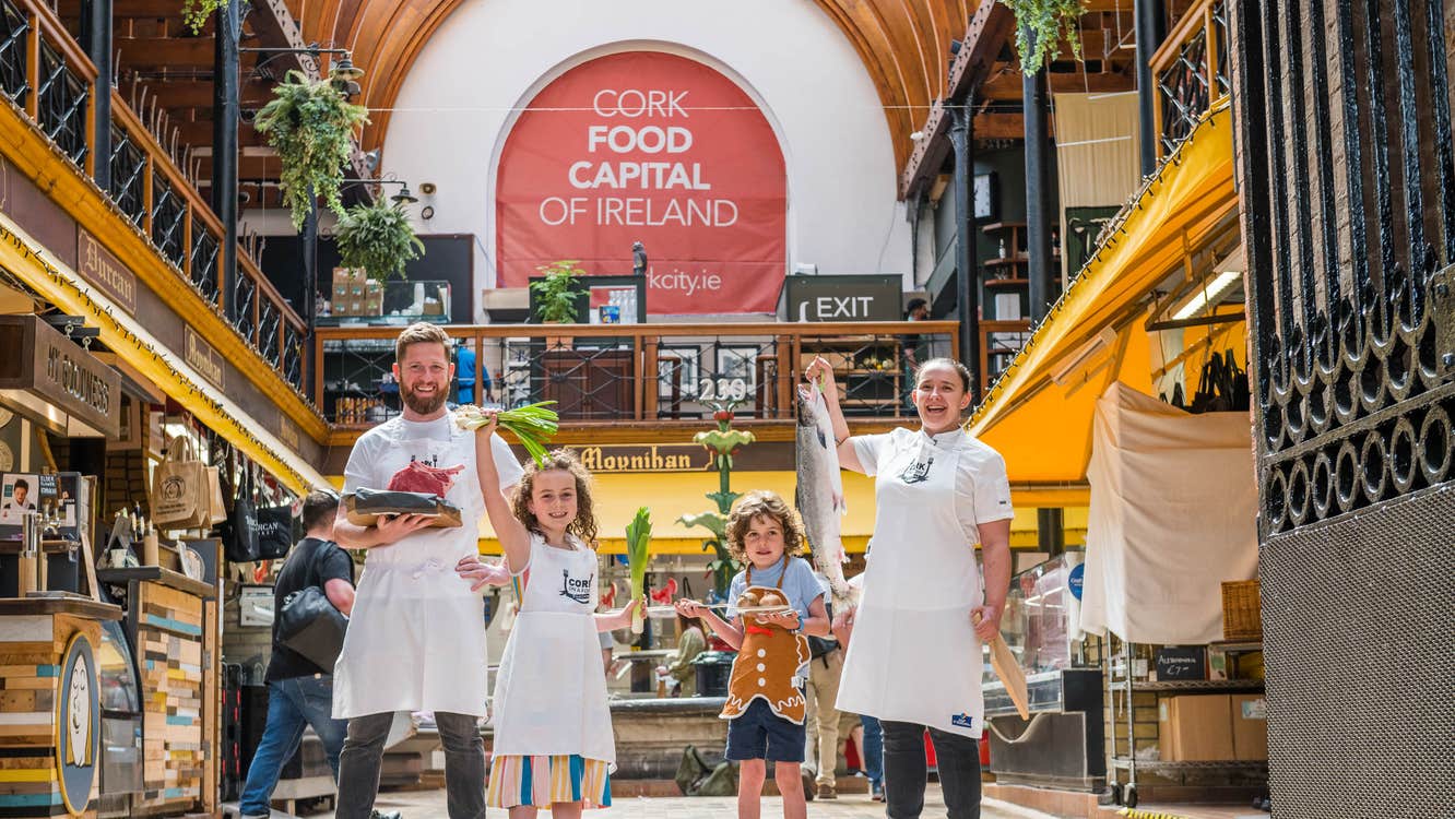 Group standing inside a market hall. One adult holds a tray of raw meat, a child holds green onions, another child holds baked goods, and one adult holds a large fish. A banner reading “CORK FOOD CAPITAL OF IRELAND” hangs above.