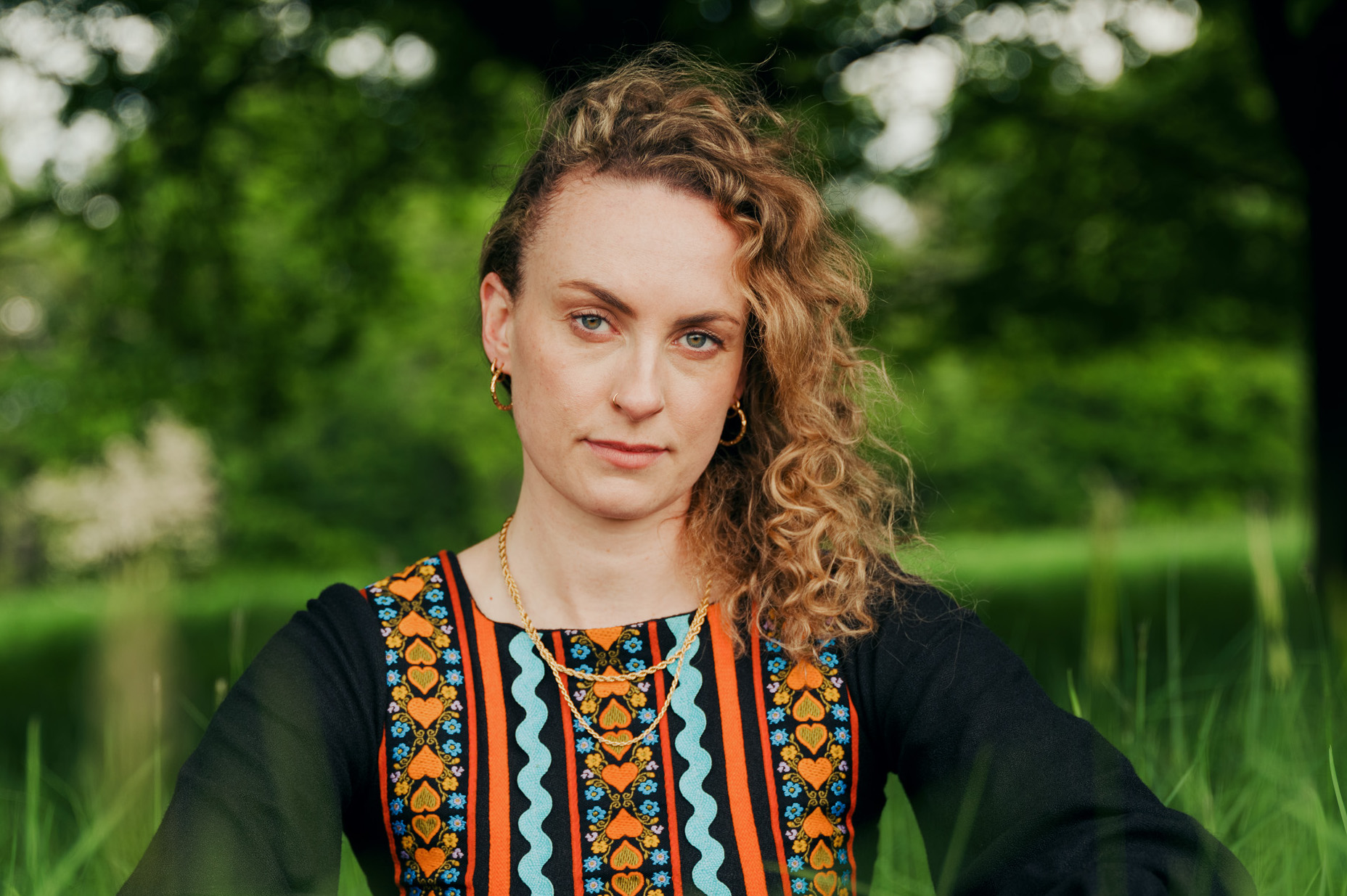 A woman in black dress with orange and blue folk pattern on is looking seriously at camera in grass with blurred greenery behind.