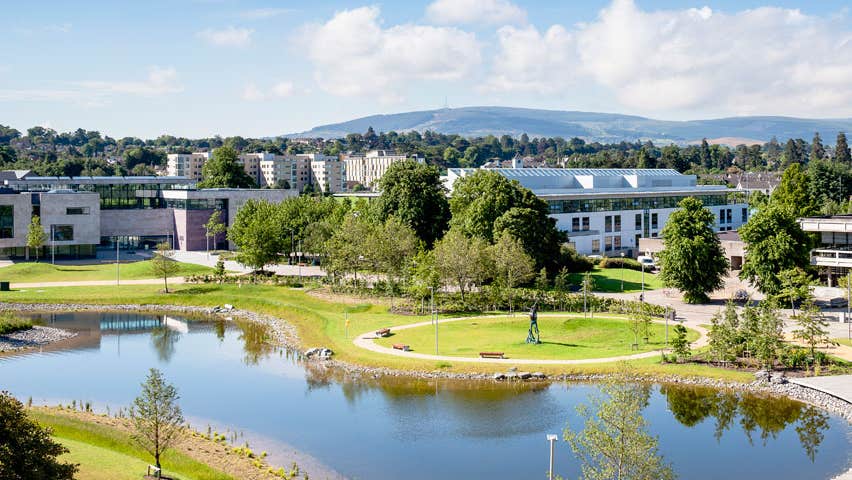 The grounds of University College Dublin with large buildings overlooking a pond and trees