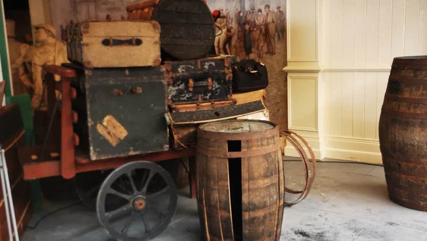 A heritage display of old luggage on a cart ready for loading on a ship with two barrels nearby