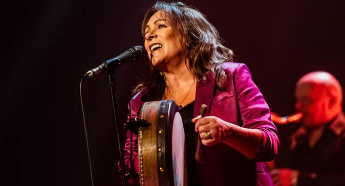 A woman is playing a hand drum, singing into a mic.
