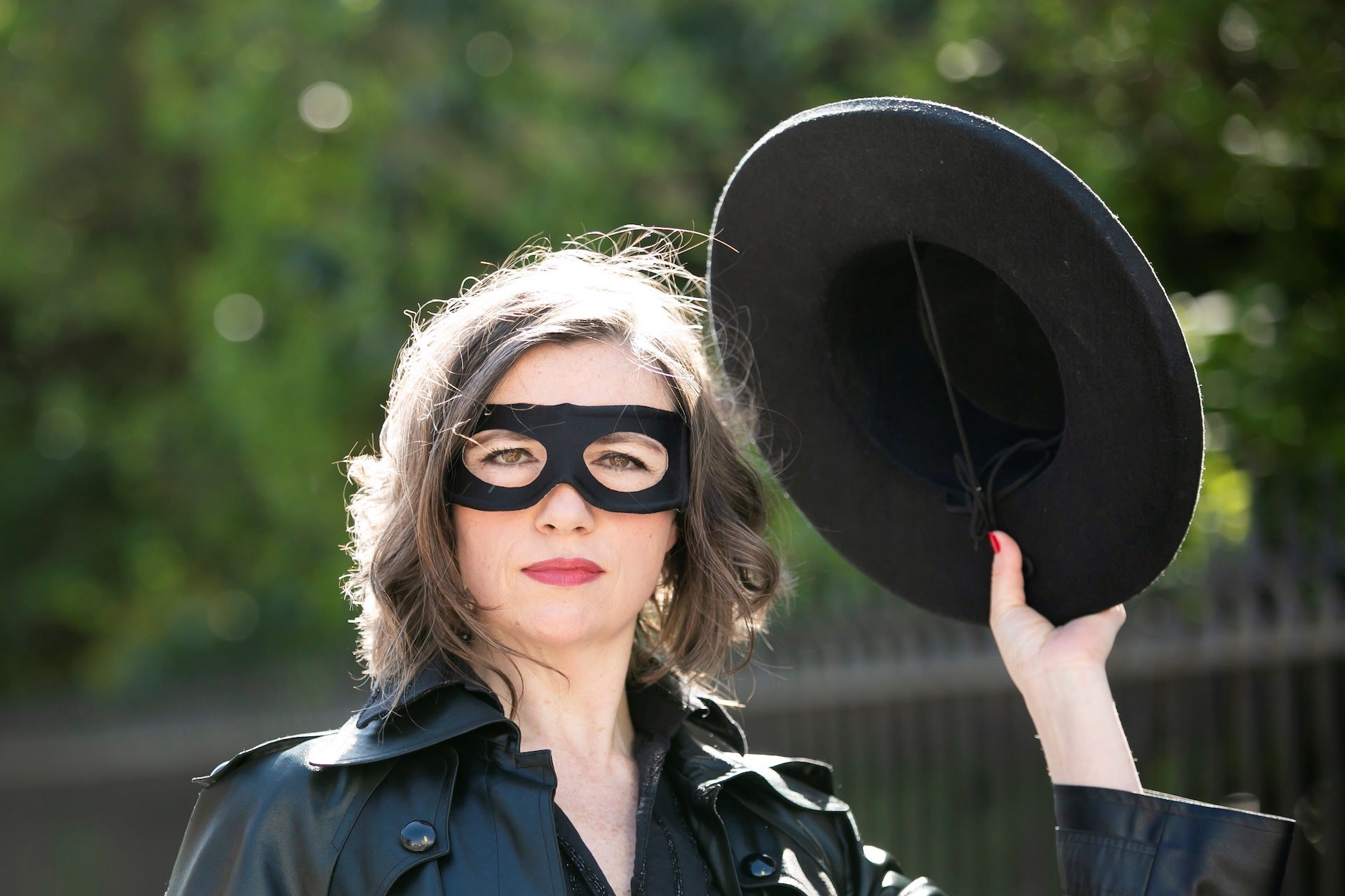 A woman outdoors dressed in black with black eye mask on, holding up a black hat in her left hand.