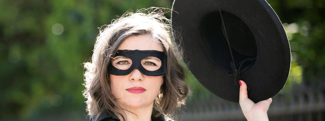 A woman outdoors dressed in black with black eye mask on, holding up a black hat in her left hand.