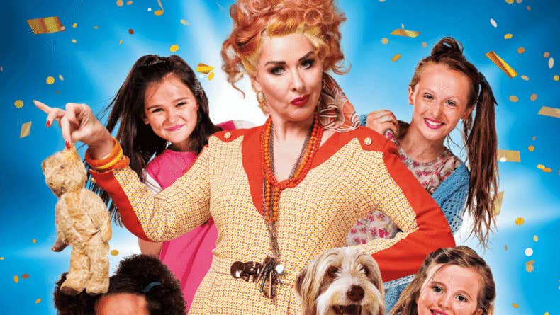 Claire Sweeney as Miss Hannigan in Annie, a woman with rye expression surrounded by 4 young girls and a dog.