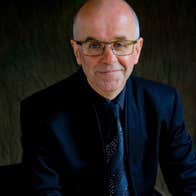 A smiling man in dark suit, tie and jacket against a dark, plain background.