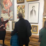 A woman is standing in front of a wall of art picture, pointing at one to small group of people in front of her standing, listening.