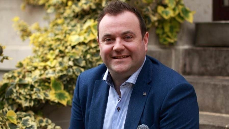 A smiling man in navy jacket seated on stone steps outside with bushes beside