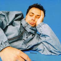Scottish jazz trailblazer corto.alto (Liam Shortall) A person in denim jacket zipped up to head is laying on blue background with head resting on left hand with eyes closed.