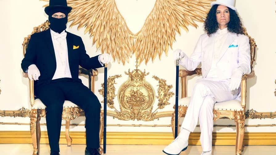 2 men seated in ornate gold chairs, one in black suit with face covered by a mask, the other in a white suit, against ornate white and old decorated wall.