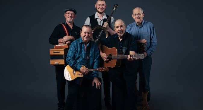 Group of 5 smiling musicians with their instruments in a tight group, against plain dark background.