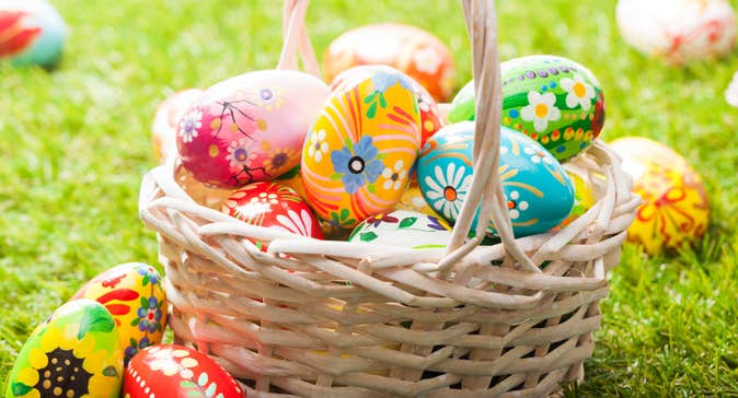 Picture of an wicker basket with tall handle containing colourful, decorated eggs with patterns and flowers, other eggs around the basket, all on grass.