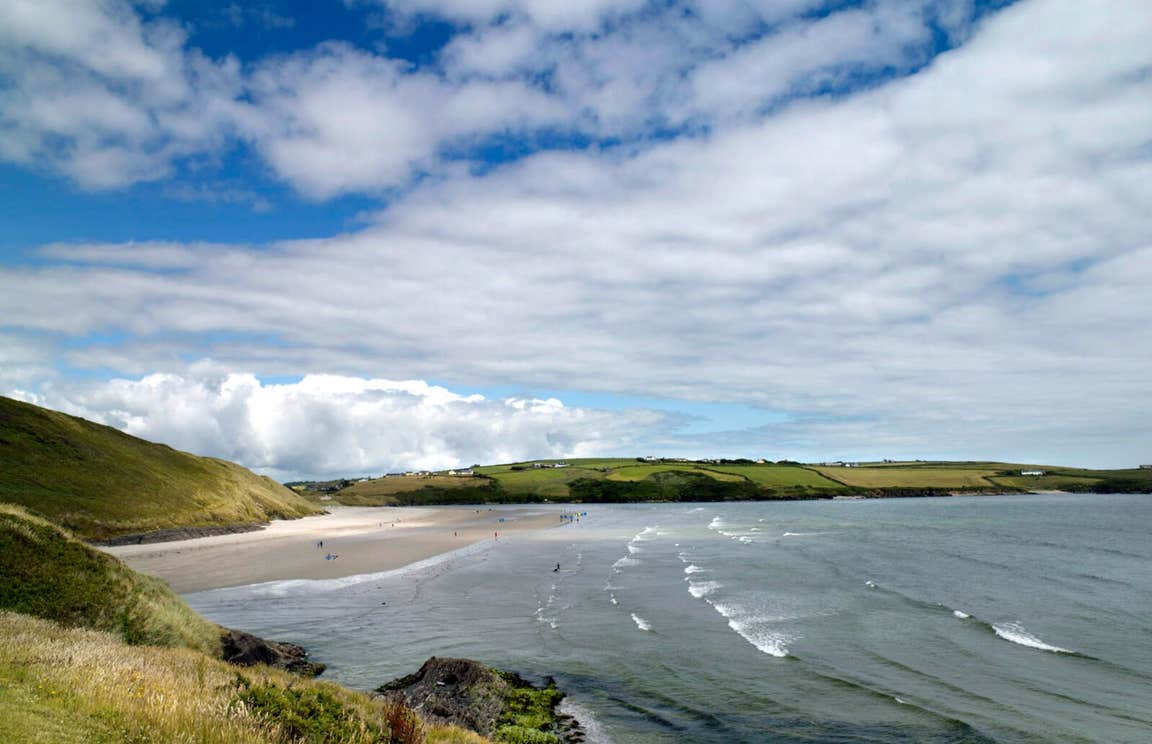 View of Inchydoney Strand with bright green grass, blue sky with some clouds, and the Atlantic ocean lapping at the sand.