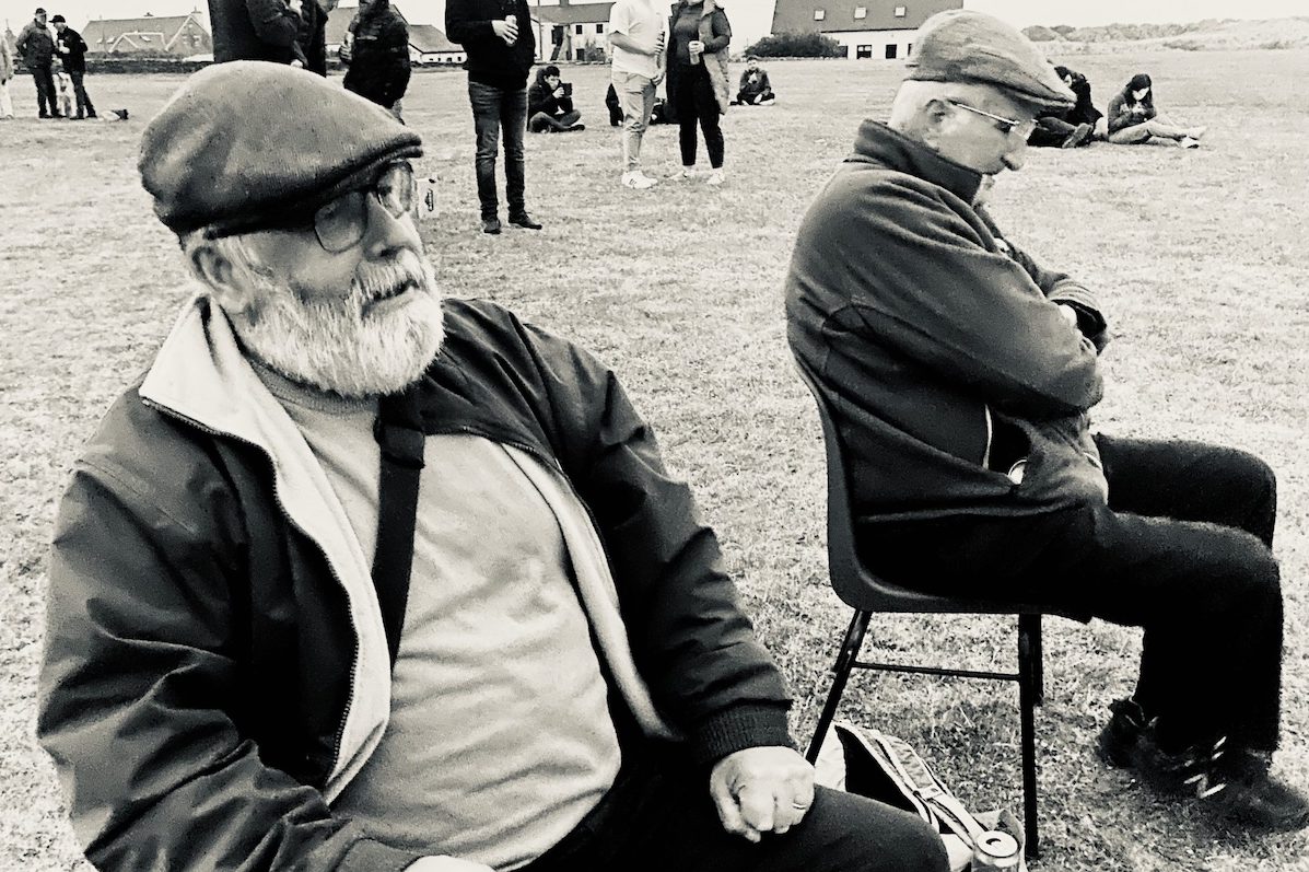 Black and white photo of 2 gentlemen, wearing flat caps, seated on plastic chairs outside on grass