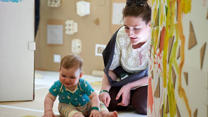 A smiling young baby seated on the floor looking down at something in front of them with an adult seated behind them.