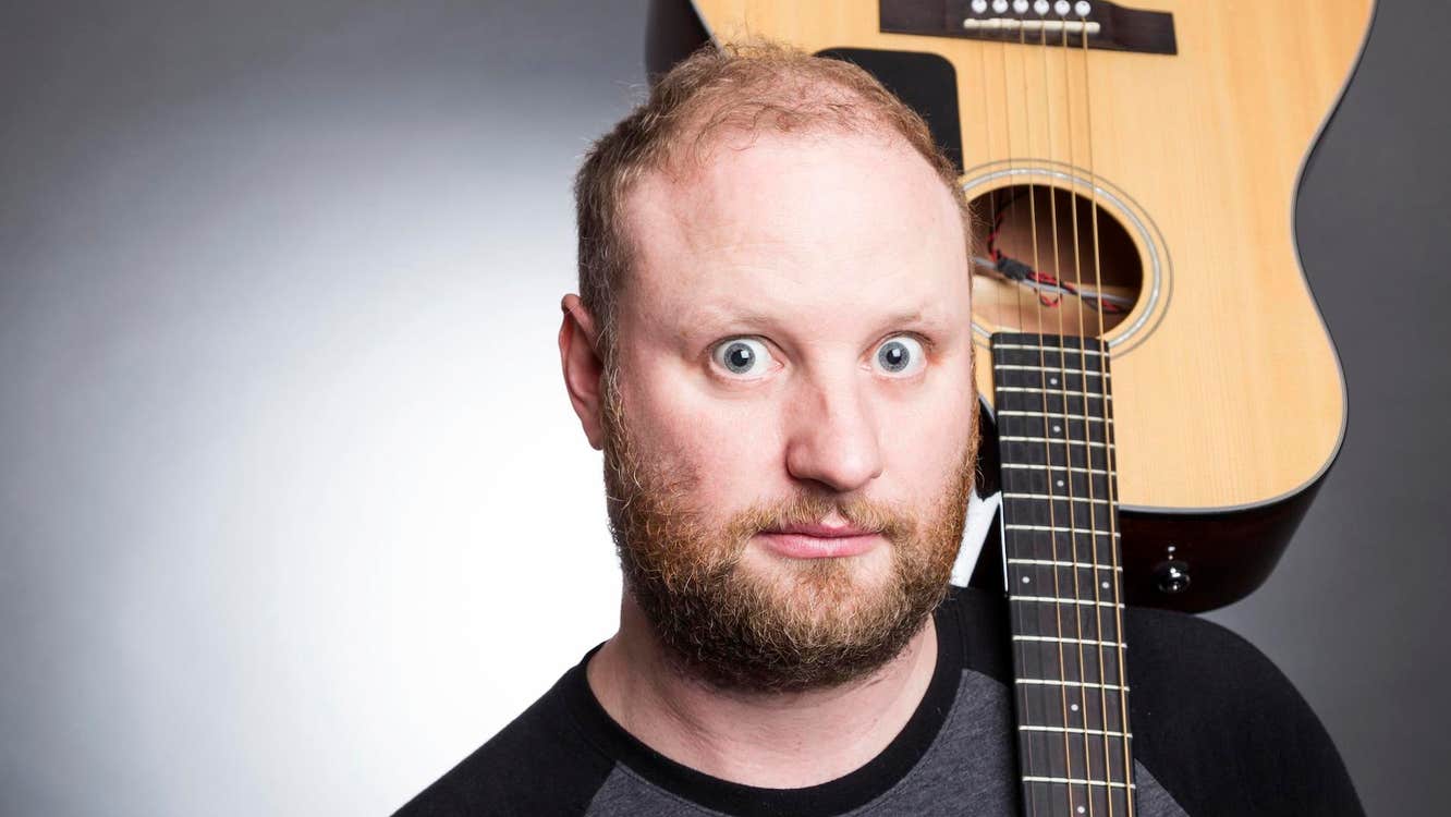 A wide eyed man is holding a guitar upside down resting on his left shoulder