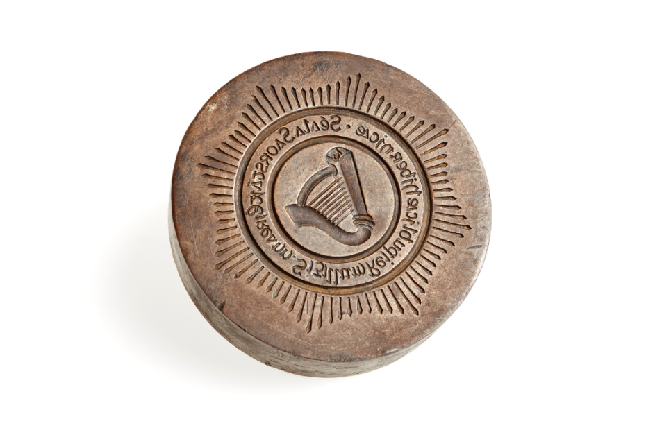 Great Seal of Saorstát Eireann, a small, brown round wooden seal with writing and harp on, against plain white background.