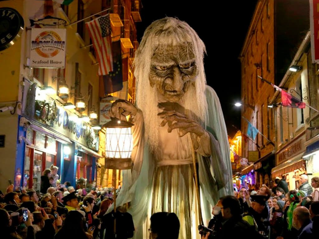 A night time parade in a narrow town street with crowds either side looking at a huge puppet of an old witch type holding a big lit lantern with flowing clothes and long white hair.