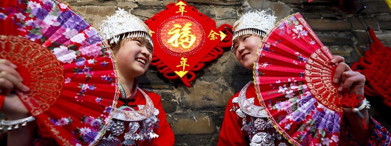 2 people in red national costumes holding large, red fans are smiling at each other, standing in front of a stone wall decorated with red and yellow decorations.