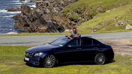 A luxury car and driver parked by a coastal road with rocky coastline in background
