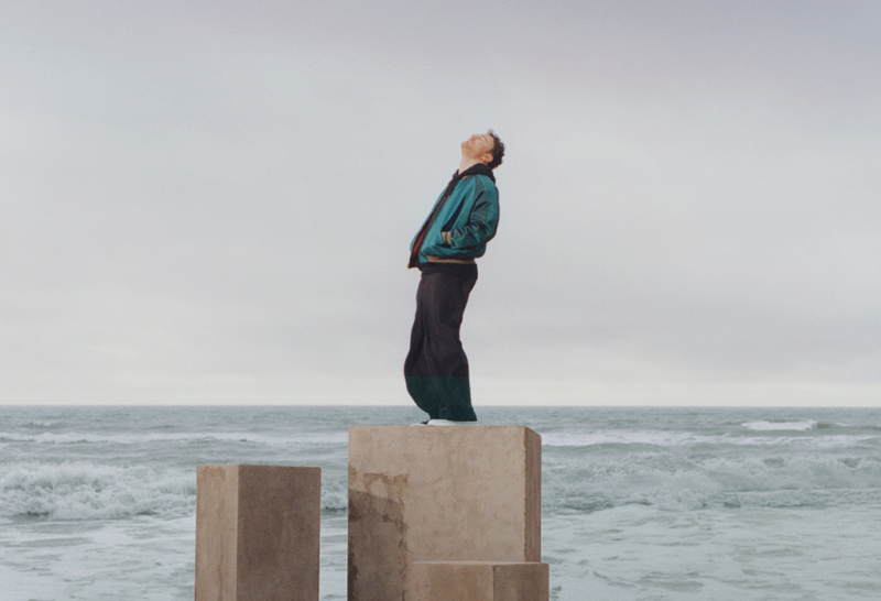 Cian Ducrot – Victory World Tour. A man standing on a large, concrete block at the edge of the sea with his hands in his pockets, with his head tilted back.