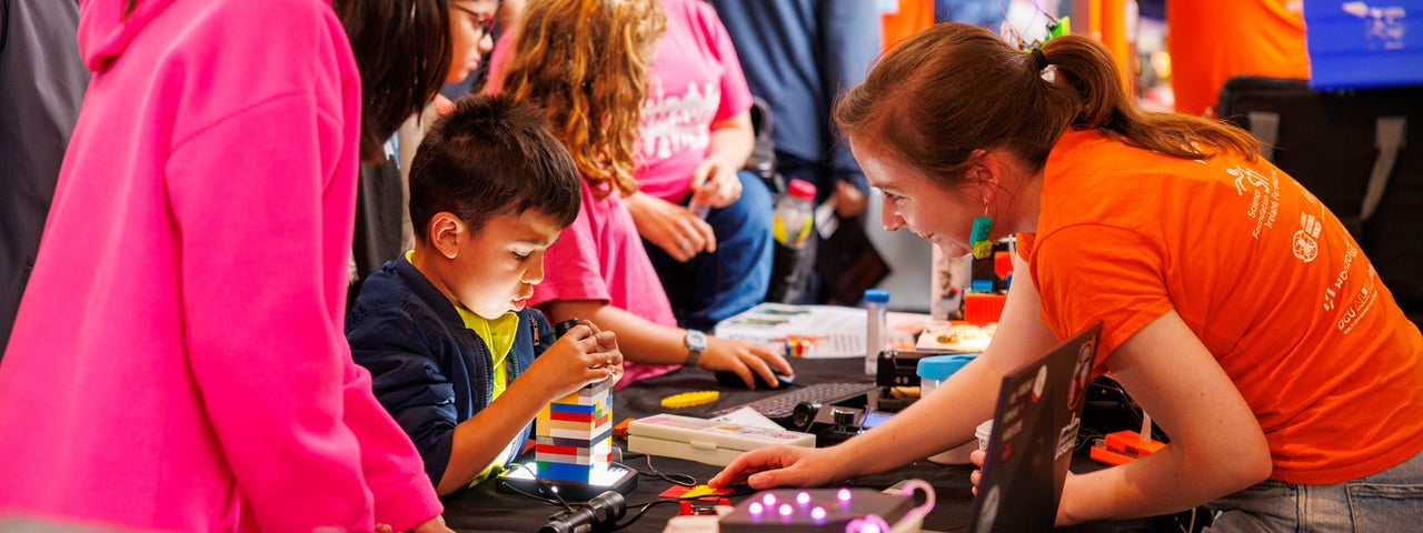 Children and adults gather around a table at a STEM workshop, exploring hands-on activities using lego, microscopes and a laptop.