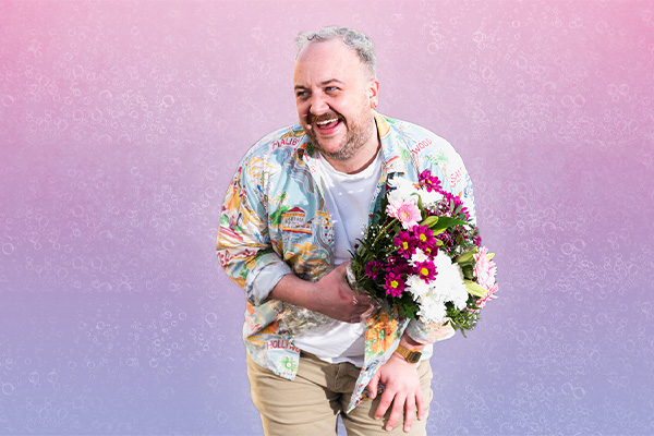 SHANE DANIEL BYRNE: BUT HE'S GAY.... A man in a brightly coloured patterned shirt is holding a bunch of flowers, laughing and looking away to his right, slightly bending forward, against plain purple/pink background.