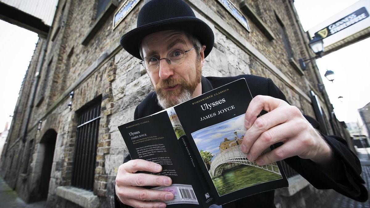 A man in black suit and hat is holding open a book in front of a corner building