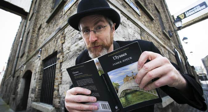 A man in black suit and hat is holding open a book in front of a corner building