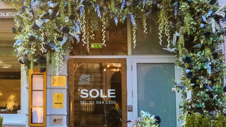The exterior of the restaurant with a flower arch over the door and a decorative yellow bicycle leaning against a branded barrel