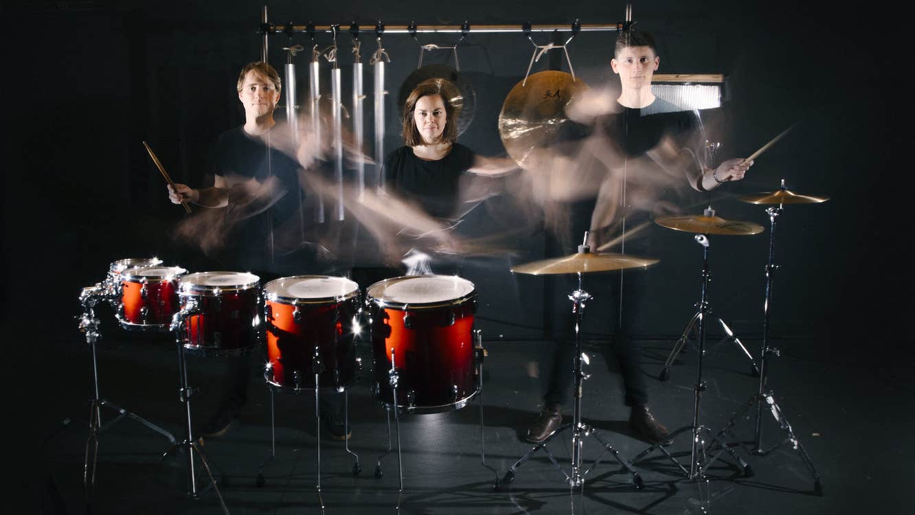 3 people standing behind drums and cymbals on standing with various percussion instruments, their hands blurred with movement