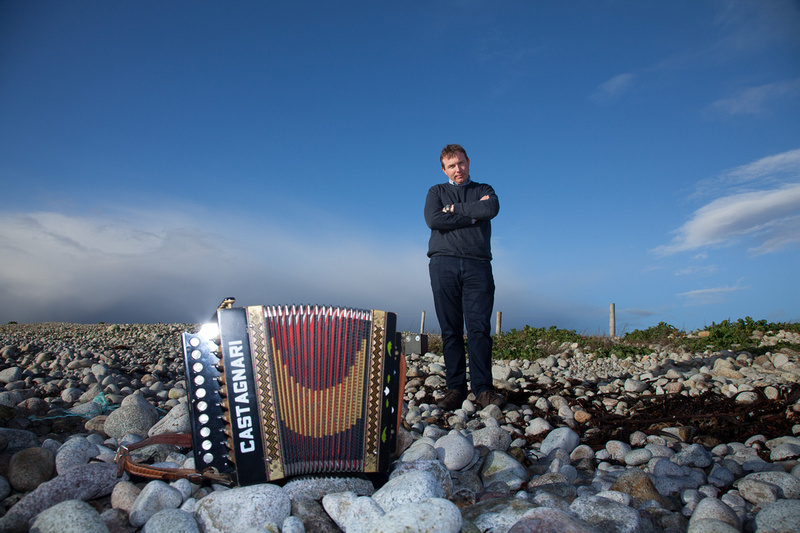 A man is stood with his arms crossed, on a rocky beach with an accordion on the ground in front of him, against a bright blue sky