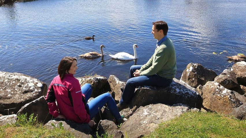 Two people sitting on rocks beside water with swans beside them